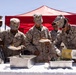 A group of U.S. Marines in desert camouflage uniforms are serving food at an outdoor event, likely a barbecue or unit gathering. Several personnel are working a food service table with metal serving trays, with one Marine pouring potato salad from a store-bought container. A red canopy tent is visible in the background.