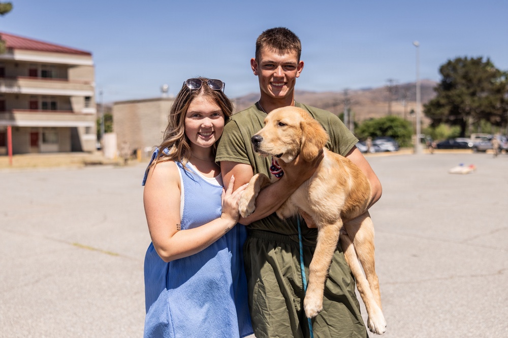 Two young adults pose together outdoors in what appears to be a military base parking lot, smiling at the camera. The male is holding a golden retriever puppy while wearing olive green military-style clothing. The setting includes institutional buildings and parked vehicles in the background under a clear blue sky.