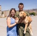 Two young adults pose together outdoors in what appears to be a military base parking lot, smiling at the camera. The male is holding a golden retriever puppy while wearing olive green military-style clothing. The setting includes institutional buildings and parked vehicles in the background under a clear blue sky.