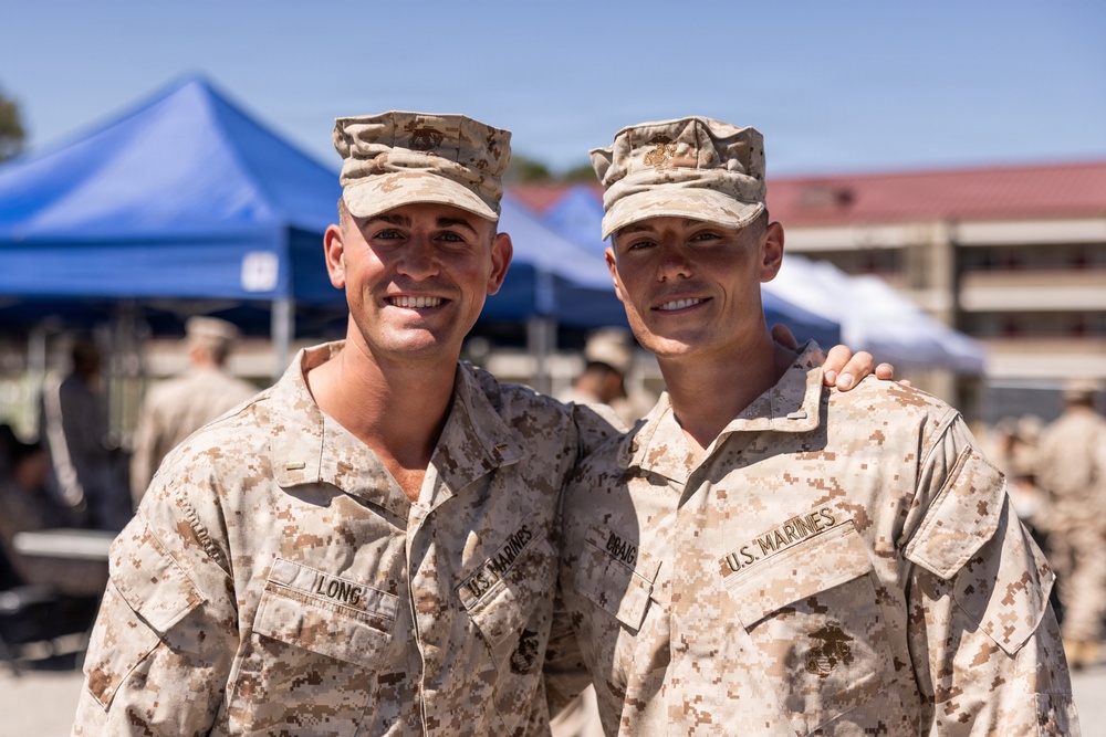 Two U.S. Marines in desert MARPAT camouflage uniforms pose together smiling at what appears to be an outdoor military event or gathering. The Marine on the left has a name tape reading 'LONG' and the one on the right appears to have a name tape reading 'BBANG' or similar. Both wear the Eagle, Globe, and Anchor emblem on their covers and chest insignia.