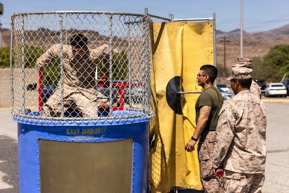Military personnel, appearing to be U.S. Marines based on their camouflage uniforms, are participating in a recreational dunk tank activity. One service member in camouflage uniform is seated inside the 'Easy Dunker 2' dunk tank, while two others stand nearby observing. The setting appears to be an outdoor military base event, likely a morale or unit celebration activity.