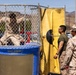 Military personnel, appearing to be U.S. Marines based on their camouflage uniforms, are participating in a recreational dunk tank activity. One service member in camouflage uniform is seated inside the 'Easy Dunker 2' dunk tank, while two others stand nearby observing. The setting appears to be an outdoor military base event, likely a morale or unit celebration activity.