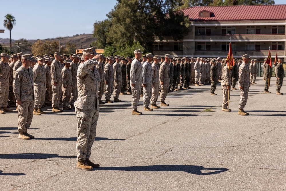 A large formation of U.S. Marines in desert MARPAT camouflage uniforms are assembled on a parade deck during what appears to be a formal military ceremony. A senior Marine in the foreground is rendering a salute to the assembled formation. Red and gold guidons are visible on the right side, consistent with U.S. Marine Corps unit colors.
