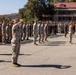A large formation of U.S. Marines in desert MARPAT camouflage uniforms are assembled on a parade deck during what appears to be a formal military ceremony. A senior Marine in the foreground is rendering a salute to the assembled formation. Red and gold guidons are visible on the right side, consistent with U.S. Marine Corps unit colors.