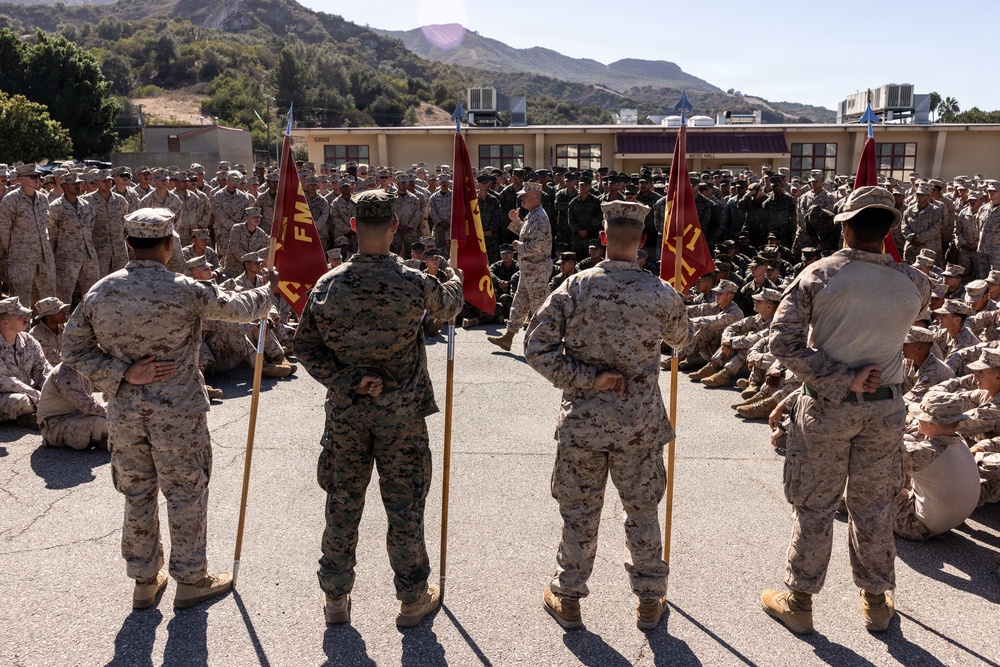 A large formation of U.S. Marines gathered in an outdoor courtyard at what appears to be a military installation, likely Camp Pendleton based on the terrain. Several Marines in the foreground are holding red guidon flags while a senior officer addresses the assembled troops. The setting suggests a formal all-hands formation, briefing, or command event.