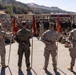 A large formation of U.S. Marines gathered in an outdoor courtyard at what appears to be a military installation, likely Camp Pendleton based on the terrain. Several Marines in the foreground are holding red guidon flags while a senior officer addresses the assembled troops. The setting suggests a formal all-hands formation, briefing, or command event.