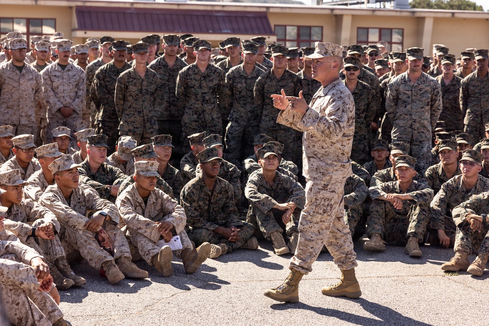 A senior military officer in U.S. Marine Corps MARPAT desert/woodland camouflage uniform addresses a large formation of Marines gathered outdoors on a paved area. The speaker is standing and gesturing while the assembled Marines are seated on the ground in front and standing in rows behind, all attentively listening. The setting appears to be a military installation with a building visible in the background.
