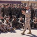 A senior military officer in U.S. Marine Corps MARPAT desert/woodland camouflage uniform addresses a large formation of Marines gathered outdoors on a paved area. The speaker is standing and gesturing while the assembled Marines are seated on the ground in front and standing in rows behind, all attentively listening. The setting appears to be a military installation with a building visible in the background.