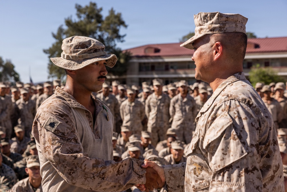 Two U.S. Marines in desert MARPAT camouflage uniforms are shaking hands in what appears to be a formal military ceremony or award presentation. A large formation of Marines in matching uniforms is assembled in the background, observing the event. The setting appears to be a military installation with a building visible behind the formation.