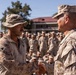 Two U.S. Marines in desert MARPAT camouflage uniforms are shaking hands in what appears to be a formal military ceremony or award presentation. A large formation of Marines in matching uniforms is assembled in the background, observing the event. The setting appears to be a military installation with a building visible behind the formation.