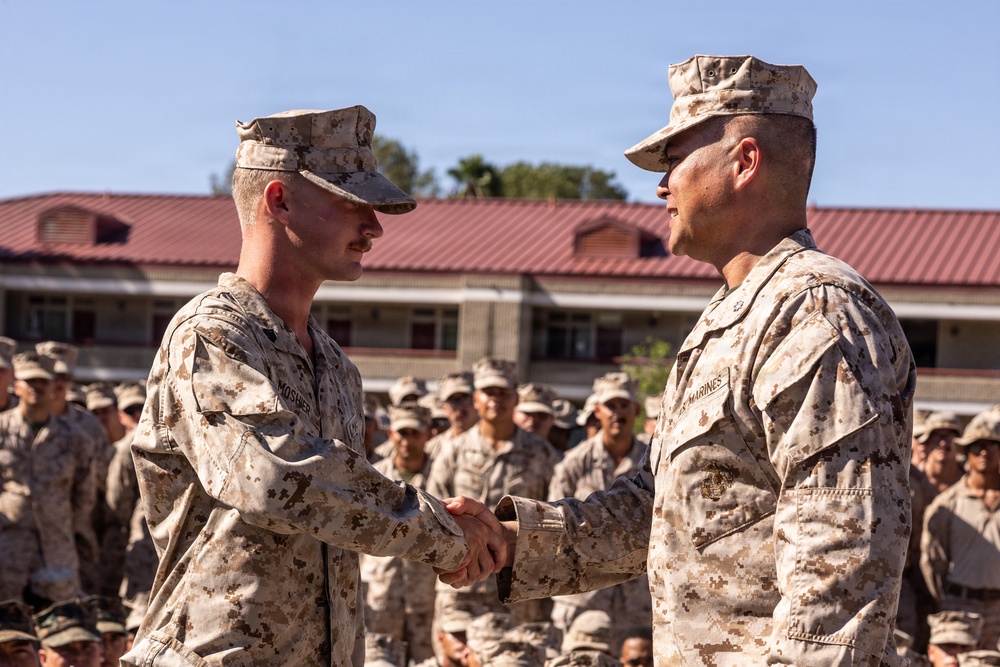 Two U.S. Marines in desert MARPAT camouflage uniforms are shaking hands in what appears to be a formal military ceremony or recognition event. A large formation of Marines stands at attention in the background, suggesting this is an official outdoor ceremony. The setting appears to be a military installation with a building featuring a red roof visible behind the formation.