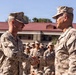 Two U.S. Marines in desert MARPAT camouflage uniforms are shaking hands in what appears to be a formal military ceremony or recognition event. A large formation of Marines stands at attention in the background, suggesting this is an official outdoor ceremony. The setting appears to be a military installation with a building featuring a red roof visible behind the formation.