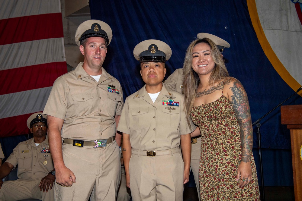 A formal U.S. Navy ceremony is taking place, likely a promotion or advancement ceremony, with personnel in khaki service uniforms posing for a photo. Three individuals stand together in the foreground — two in Navy Chief Petty Officer uniforms and one civilian woman in a floral dress with visible tattoos. An American flag and blue ceremonial backdrop are visible in the background.