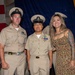 A formal U.S. Navy ceremony is taking place, likely a promotion or advancement ceremony, with personnel in khaki service uniforms posing for a photo. Three individuals stand together in the foreground — two in Navy Chief Petty Officer uniforms and one civilian woman in a floral dress with visible tattoos. An American flag and blue ceremonial backdrop are visible in the background.