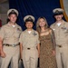 Four individuals pose for a photo at what appears to be a U.S. Navy ceremony aboard the USS Nimitz (CVN-68). Three are wearing U.S. Navy Service Khaki uniforms with chief petty officer covers and ribbons, while one civilian woman in a floral dress stands among them. The setting includes an American flag, a blue ceremonial backdrop, and the USS Nimitz podium seal.