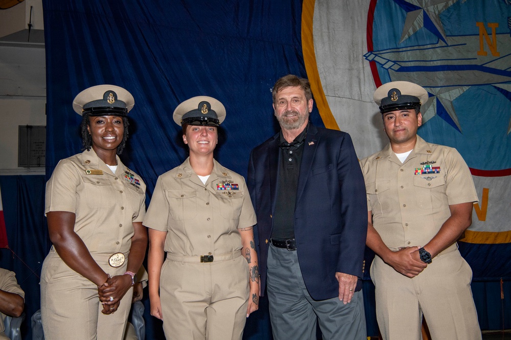 Three U.S. Navy personnel in khaki service uniforms pose with a civilian man in a navy blazer at what appears to be a formal military ceremony or recognition event. The backdrop features a large blue ceremonial banner with a Navy insignia. The three sailors wear chief petty officer covers (combination caps) and display various service ribbons and badges.