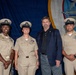 Three U.S. Navy personnel in khaki service uniforms pose with a civilian man in a navy blazer at what appears to be a formal military ceremony or recognition event. The backdrop features a large blue ceremonial banner with a Navy insignia. The three sailors wear chief petty officer covers (combination caps) and display various service ribbons and badges.