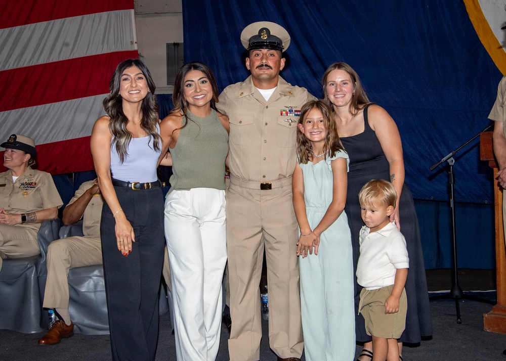A U.S. Navy chief petty officer in dress khaki uniform poses with what appears to be family members during a military ceremony or promotion event. The group includes two adult women, a young girl, and a toddler boy, all dressed in celebratory attire. An American flag and navy blue draping are visible in the background, along with other uniformed personnel seated nearby.