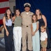 A U.S. Navy chief petty officer in dress khaki uniform poses with what appears to be family members during a military ceremony or promotion event. The group includes two adult women, a young girl, and a toddler boy, all dressed in celebratory attire. An American flag and navy blue draping are visible in the background, along with other uniformed personnel seated nearby.