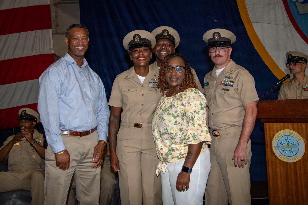 A group photo taken aboard the USS Nimitz (CVN 68), visible on the podium seal in the background. Three U.S. Navy personnel in khaki chief petty officer uniforms pose with two civilians — a man in a blue dress shirt and a woman in a floral top — in front of an American flag and blue ceremonial backdrop. The setting appears to be a formal military ceremony or promotion event.