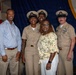 A group photo taken aboard the USS Nimitz (CVN 68), visible on the podium seal in the background. Three U.S. Navy personnel in khaki chief petty officer uniforms pose with two civilians — a man in a blue dress shirt and a woman in a floral top — in front of an American flag and blue ceremonial backdrop. The setting appears to be a formal military ceremony or promotion event.