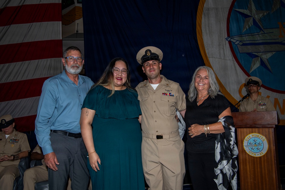 A U.S. Navy service member in dress khaki uniform poses with three civilians at what appears to be a military promotion or recognition ceremony. The group of four stands in front of an American flag and a Navy command insignia backdrop. A podium bearing a Navy seal is visible to the right, and additional uniformed personnel are present in the background.