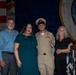A U.S. Navy service member in dress khaki uniform poses with three civilians at what appears to be a military promotion or recognition ceremony. The group of four stands in front of an American flag and a Navy command insignia backdrop. A podium bearing a Navy seal is visible to the right, and additional uniformed personnel are present in the background.