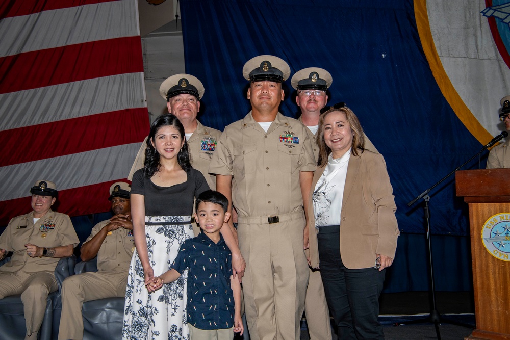 A U.S. Navy promotion or advancement ceremony taking place aboard what appears to be a naval vessel, likely the USS Nimitz based on the partially visible podium seal. A central Navy chief petty officer poses with family members including a woman, a young boy, and an older woman, flanked by two other chief petty officers in tan service uniforms. An American flag and blue ceremonial draping serve as the backdrop.