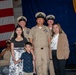 A U.S. Navy promotion or advancement ceremony taking place aboard what appears to be a naval vessel, likely the USS Nimitz based on the partially visible podium seal. A central Navy chief petty officer poses with family members including a woman, a young boy, and an older woman, flanked by two other chief petty officers in tan service uniforms. An American flag and blue ceremonial draping serve as the backdrop.