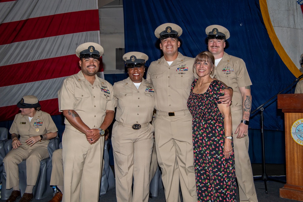 A group photo at what appears to be a U.S. Navy ceremony or promotion event. Four Navy personnel in tan/khaki service uniforms are posing with one civilian woman in a floral dress. An American flag and blue ceremonial backdrop are visible in the background.