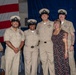 A group photo at what appears to be a U.S. Navy ceremony or promotion event. Four Navy personnel in tan/khaki service uniforms are posing with one civilian woman in a floral dress. An American flag and blue ceremonial backdrop are visible in the background.