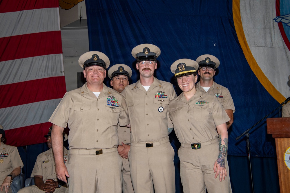 A group of five U.S. Navy personnel in tan/khaki service uniforms are posing together at what appears to be a formal military ceremony or event. They are standing in front of a blue backdrop and an American flag, with a podium visible to the right. The individuals are wearing chief petty officer or senior enlisted covers (caps) with gold insignia.