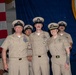 A group of five U.S. Navy personnel in tan/khaki service uniforms are posing together at what appears to be a formal military ceremony or event. They are standing in front of a blue backdrop and an American flag, with a podium visible to the right. The individuals are wearing chief petty officer or senior enlisted covers (caps) with gold insignia.