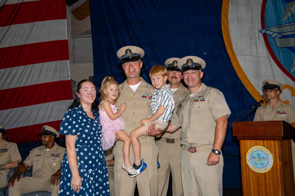 A military ceremony aboard USS Nimitz (CVN-68) featuring Navy personnel in service khaki uniforms posing for a group photo with family members, including two young children. The setting appears to be a formal event such as a promotion, retirement, or change of command ceremony. An American flag and the USS Nimitz ship's seal are visible in the background.