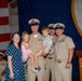 A military ceremony aboard USS Nimitz (CVN-68) featuring Navy personnel in service khaki uniforms posing for a group photo with family members, including two young children. The setting appears to be a formal event such as a promotion, retirement, or change of command ceremony. An American flag and the USS Nimitz ship's seal are visible in the background.