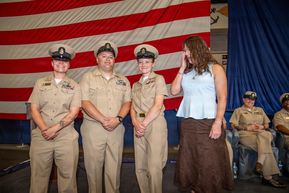 A military ceremony featuring three U.S. Navy personnel in khaki service uniforms standing in front of a large American flag. A civilian woman in a white top and brown skirt stands beside them, appearing emotional with her hand raised to her face. Additional uniformed personnel are seated in the background, suggesting a formal promotion or recognition ceremony.