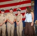 A military ceremony featuring three U.S. Navy personnel in khaki service uniforms standing in front of a large American flag. A civilian woman in a white top and brown skirt stands beside them, appearing emotional with her hand raised to her face. Additional uniformed personnel are seated in the background, suggesting a formal promotion or recognition ceremony.