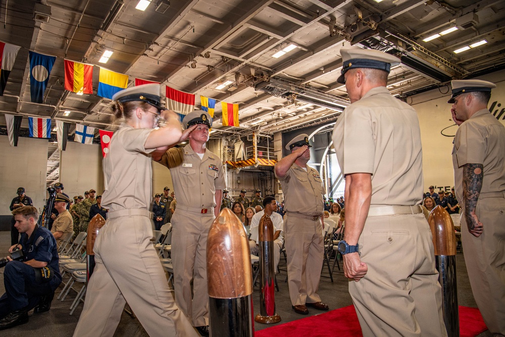 A formal U.S. Navy military ceremony is taking place inside what appears to be a ship's hangar bay, likely a change of command or promotion ceremony. Personnel in khaki service uniforms are exchanging salutes on a red carpet, flanked by ceremonial items. The space is decorated with international maritime signal flags hanging from the overhead structure, with an audience of military and civilian attendees seated in the background.