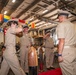 A formal U.S. Navy military ceremony is taking place inside what appears to be a ship's hangar bay, likely a change of command or promotion ceremony. Personnel in khaki service uniforms are exchanging salutes on a red carpet, flanked by ceremonial items. The space is decorated with international maritime signal flags hanging from the overhead structure, with an audience of military and civilian attendees seated in the background.