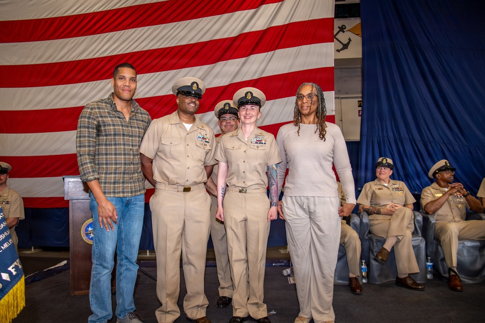 A group of four individuals pose for a photo in front of a large American flag at what appears to be a U.S. Navy ceremony or event. Two individuals are wearing U.S. Navy Service Khaki uniforms with chief petty officer covers, while two others are in civilian clothing. Additional Navy personnel are seated in the background.
