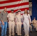 A group of four individuals pose for a photo in front of a large American flag at what appears to be a U.S. Navy ceremony or event. Two individuals are wearing U.S. Navy Service Khaki uniforms with chief petty officer covers, while two others are in civilian clothing. Additional Navy personnel are seated in the background.