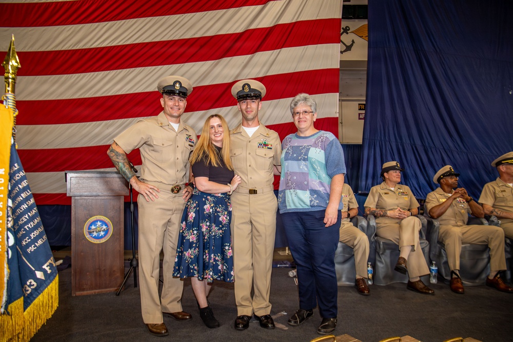 A group of four people pose for a photo at what appears to be a U.S. Navy promotion or graduation ceremony. Two individuals are wearing U.S. Navy Service Khaki uniforms with combination covers, flanking two civilian women. The event is held indoors in front of a large American flag backdrop with Navy insignia visible.