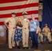 A group of four people pose for a photo at what appears to be a U.S. Navy promotion or graduation ceremony. Two individuals are wearing U.S. Navy Service Khaki uniforms with combination covers, flanking two civilian women. The event is held indoors in front of a large American flag backdrop with Navy insignia visible.