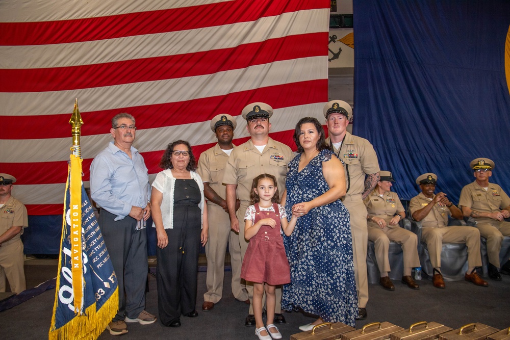 A military ceremony, likely a promotion or advancement ceremony, taking place in front of a large American flag backdrop with a U.S. Navy anchor insignia visible. Several Navy personnel in khaki uniforms are posing with civilian family members, including an older couple, a woman in a blue floral dress, and a young girl in a red pinafore. A Navy unit guidon/pennant with 'NAVIGATION' text is visible on the left side of the frame.