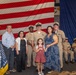 A military ceremony, likely a promotion or advancement ceremony, taking place in front of a large American flag backdrop with a U.S. Navy anchor insignia visible. Several Navy personnel in khaki uniforms are posing with civilian family members, including an older couple, a woman in a blue floral dress, and a young girl in a red pinafore. A Navy unit guidon/pennant with 'NAVIGATION' text is visible on the left side of the frame.