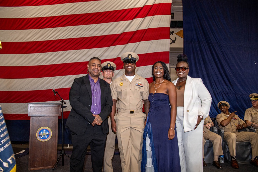 A military promotion or award ceremony is taking place in front of a large American flag backdrop. A U.S. Navy chief petty officer in service khaki uniform is posing for a photo with three civilians, likely family members. Additional uniformed Navy personnel are seated in the background observing the ceremony.