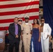 A military promotion or award ceremony is taking place in front of a large American flag backdrop. A U.S. Navy chief petty officer in service khaki uniform is posing for a photo with three civilians, likely family members. Additional uniformed Navy personnel are seated in the background observing the ceremony.