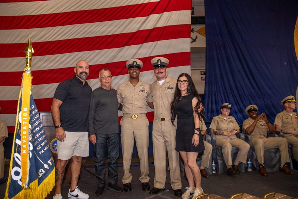 A military ceremony, likely a promotion or advancement event, is taking place aboard a U.S. Navy vessel or installation. Two uniformed Navy Chief Petty Officers pose with three civilians in front of a large American flag and blue draping. Additional uniformed Navy personnel are seated in the background observing the event.
