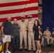 A military ceremony, likely a promotion or advancement event, is taking place aboard a U.S. Navy vessel or installation. Two uniformed Navy Chief Petty Officers pose with three civilians in front of a large American flag and blue draping. Additional uniformed Navy personnel are seated in the background observing the event.
