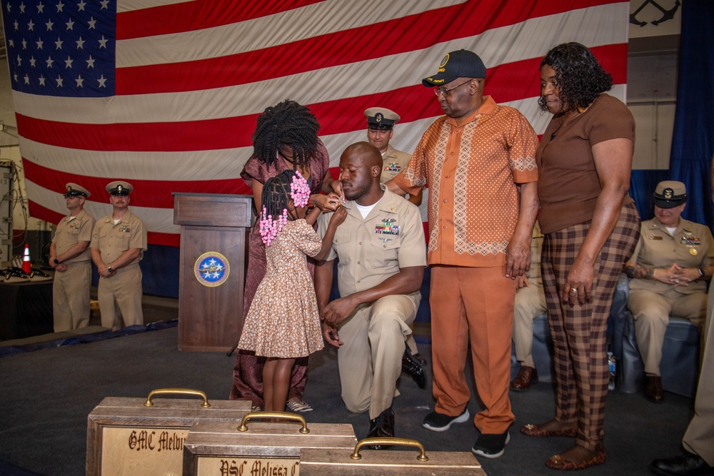 A U.S. Navy promotion or advancement ceremony is taking place in front of a large American flag. A sailor in khaki dress uniform is kneeling while a young girl pins insignia or a device onto his collar, surrounded by family members and fellow naval personnel. Engraved wooden plaques or cases are visible in the foreground, indicating this is a formal recognition event.